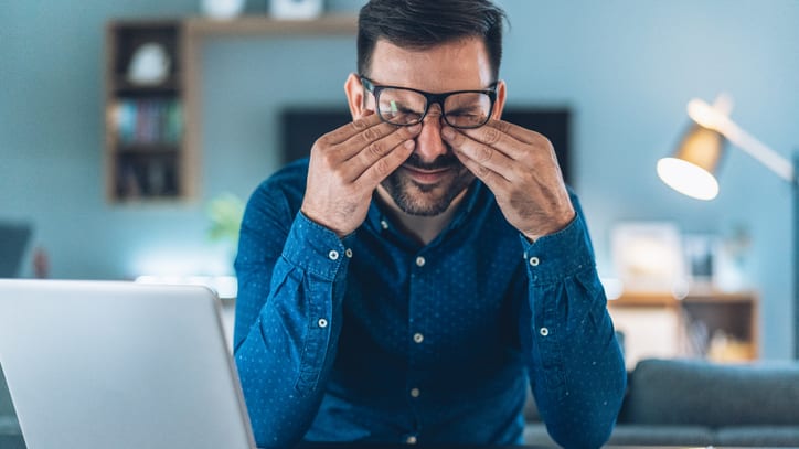 A man looking stressed rubbing his eyes while working on his laptop.