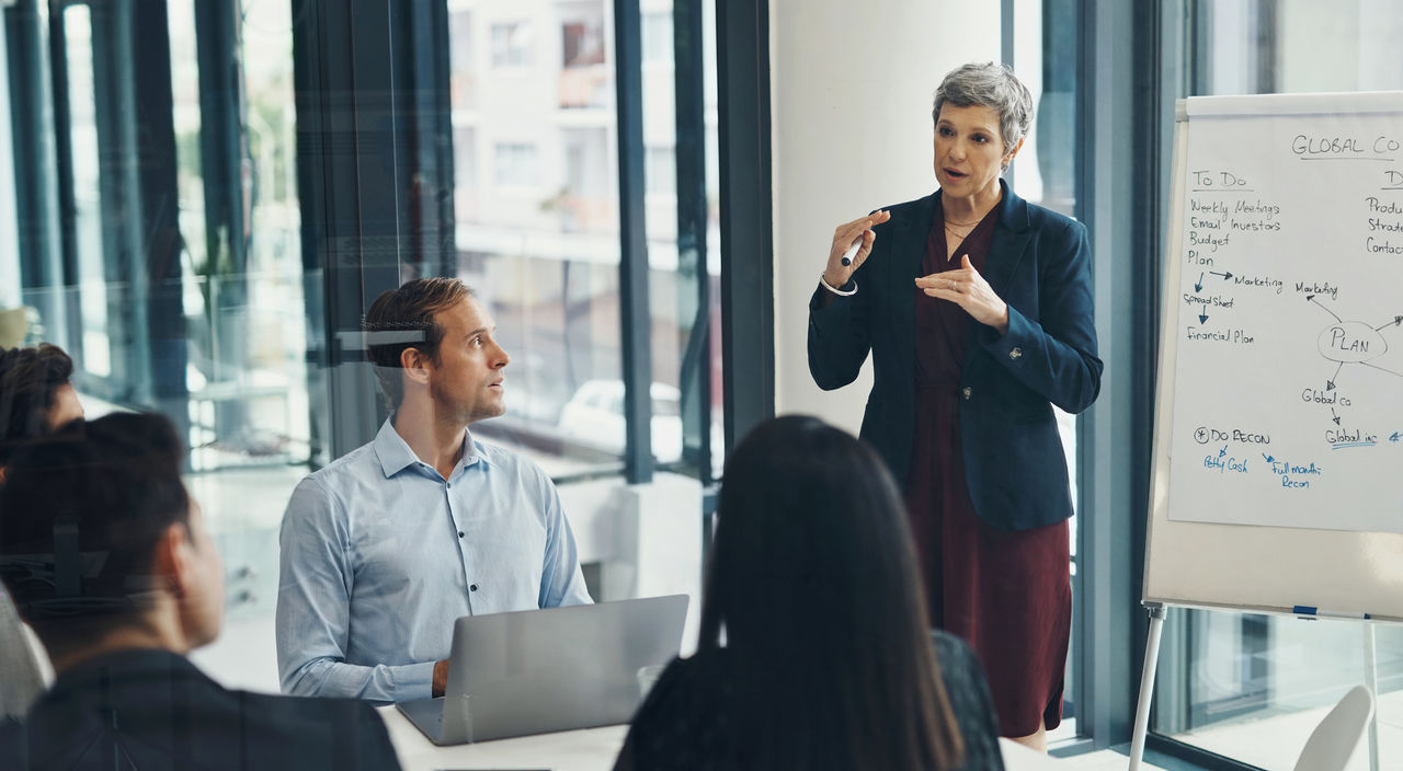 1987166734 Meeting, whiteboard and senior business woman in boardroom with team for training or coaching. Presentation, planning or strategy with manager and employee group in office for upskill development