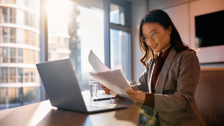 A business woman is sitting at a table with papers and a laptop.