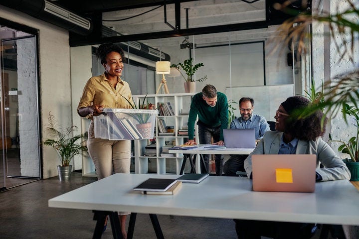 African american female worker coming back to work, holding box with office items and talking with her colleague who sitting and working on laptop while male coworkers brainstorming in the background