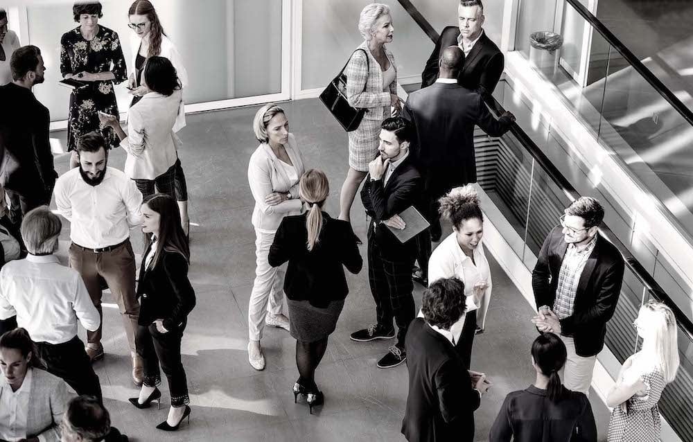 A group of people in professional clothing are seen from overhead speaking cordially in an office.