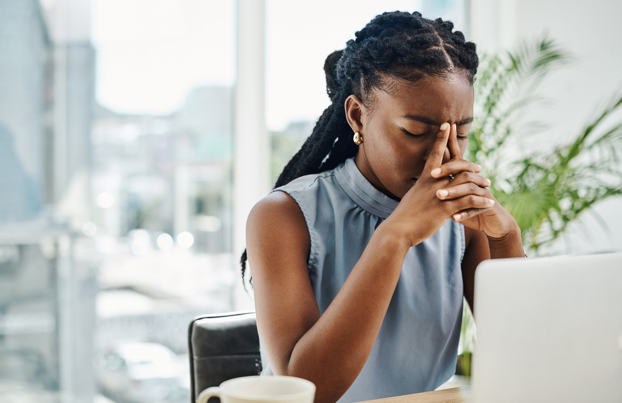 A professional woman in a blue top sits at a desk in front a computer and covers her face in frustration.