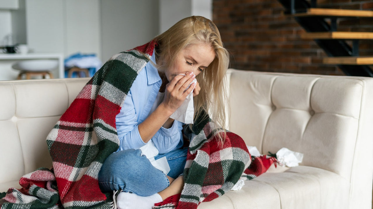 A woman sitting on a couch with a blanket over her shoulders blowing her nose.