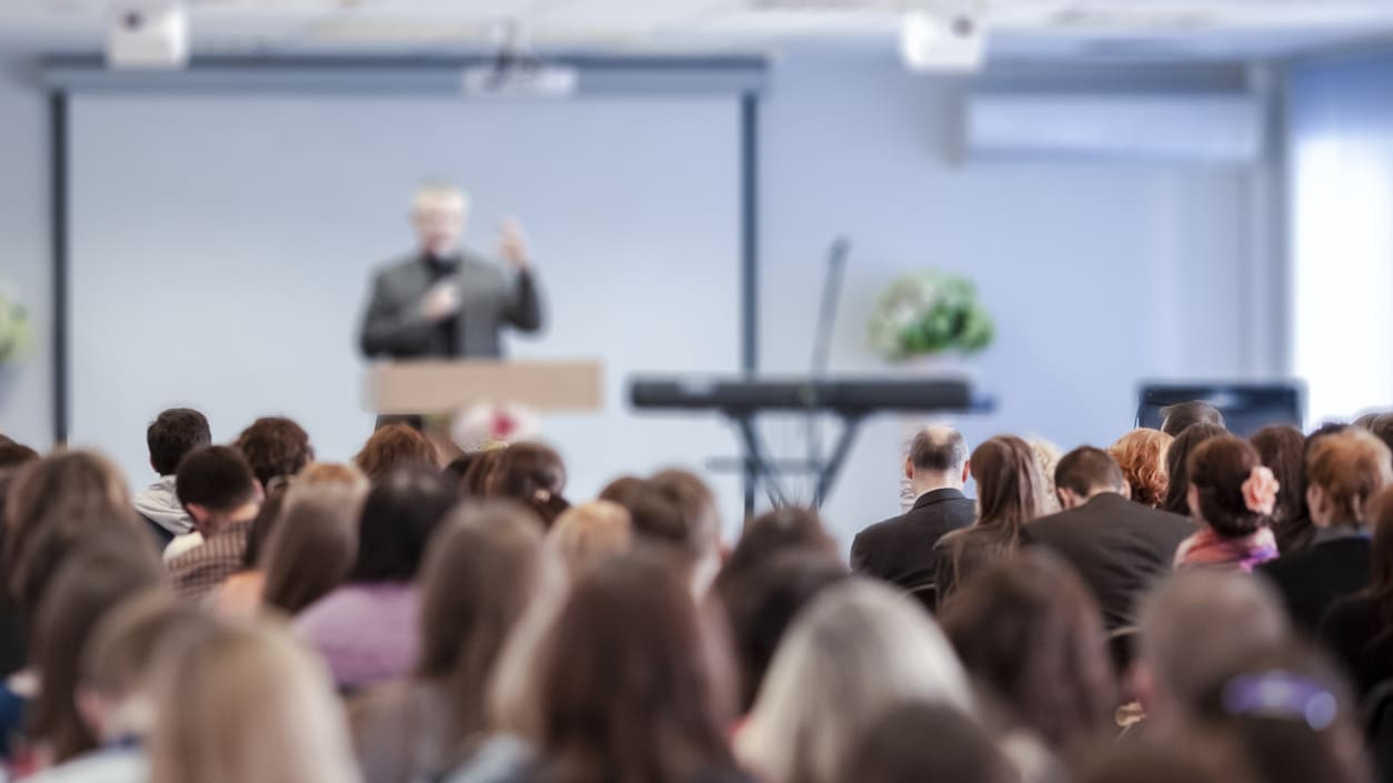 A meeting room filled with employees and the presenter gesturing at the front of the room.