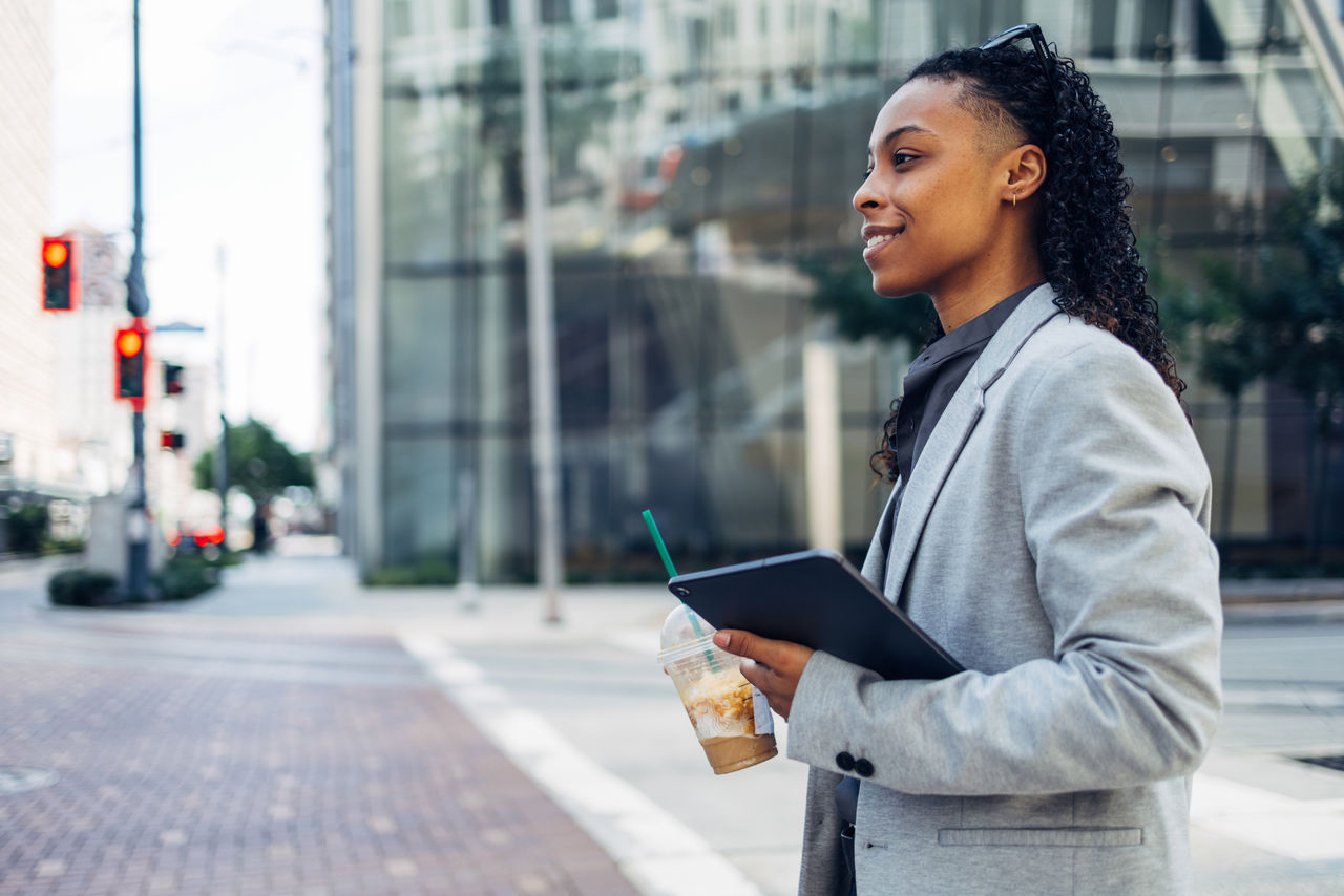 Confident businesswoman wearing a blazer, walking in the city with a coffee and tablet. Urban professional on the go.