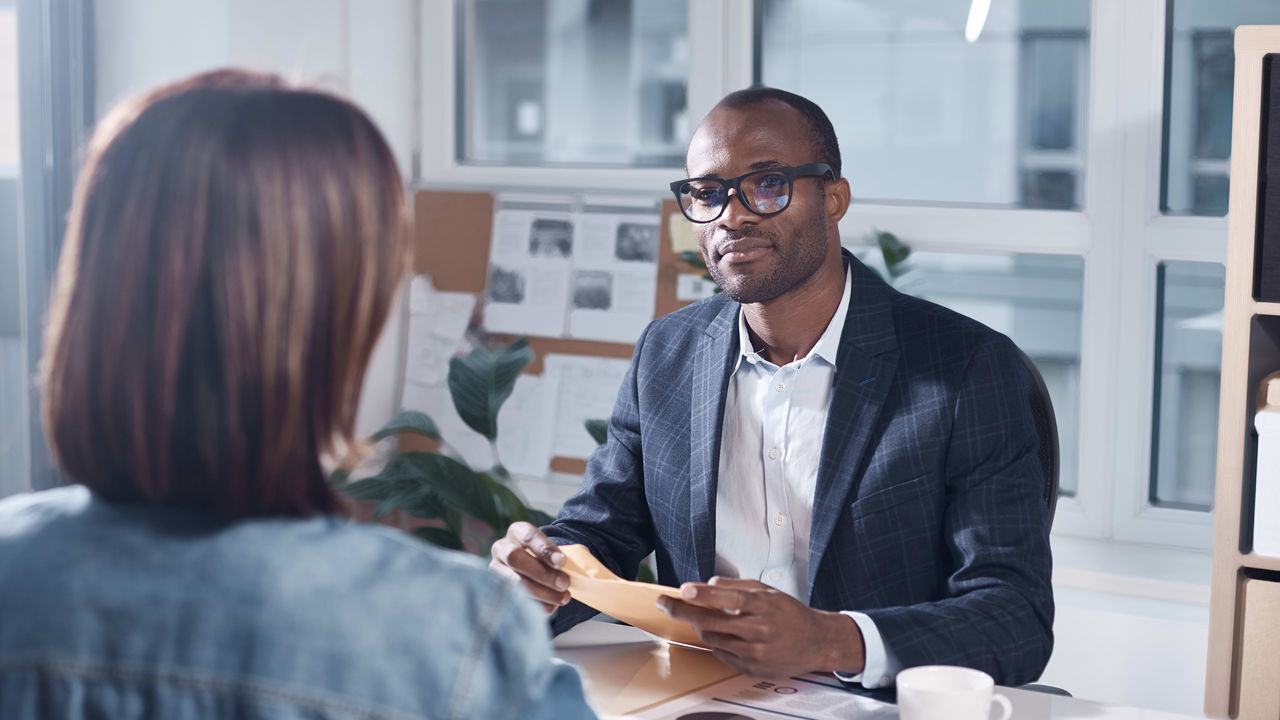 A man is talking to a woman at a desk in an office.