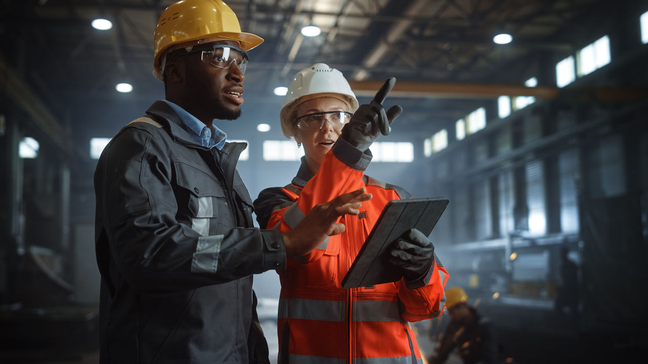 Two manufacturing employees, both with hard helmets on, on manufacturing floor, and one of them is pointing.