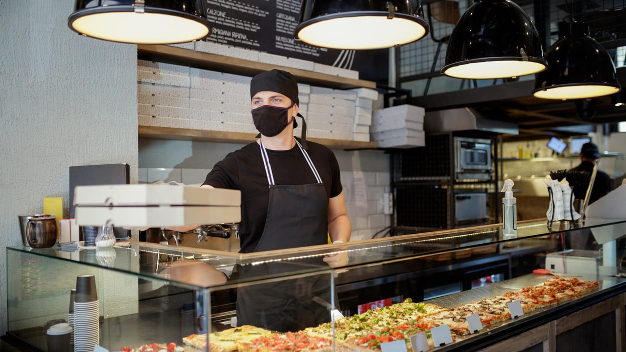 A man in a black mask is standing behind a counter full of pizzas.