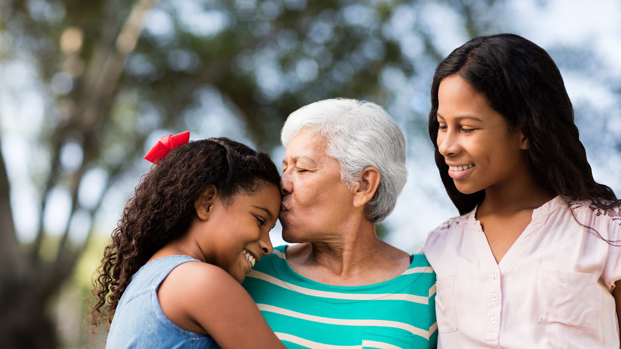 Three young girls hugging an older woman in the park.