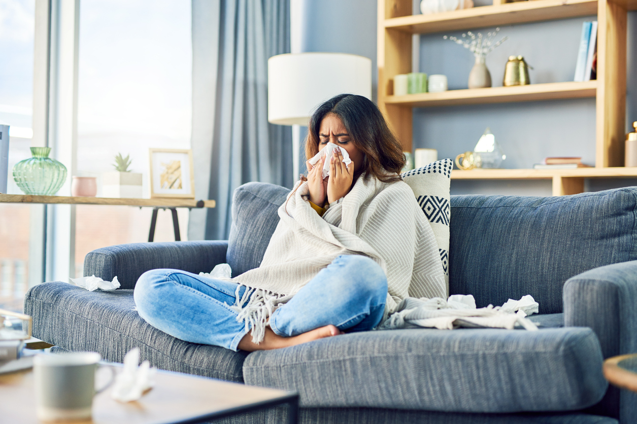A woman sneezing, wrapped up in a blanket, sitting cross-legged on a sofa.