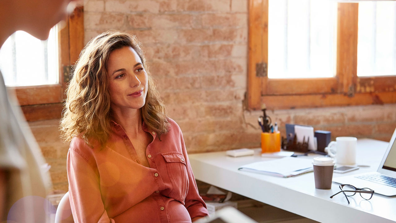 A pregnant woman is talking to another woman in an office.