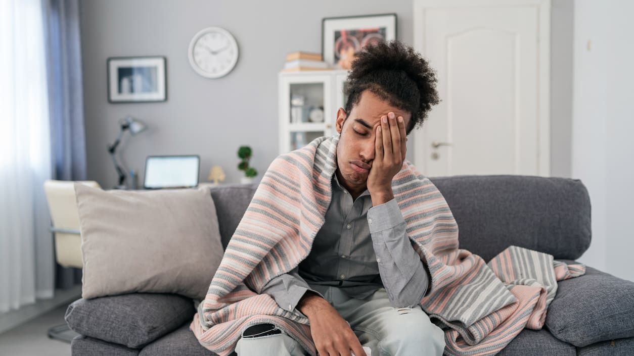 A sick worker on the couch, covering one eye and wrapped in a blanket.