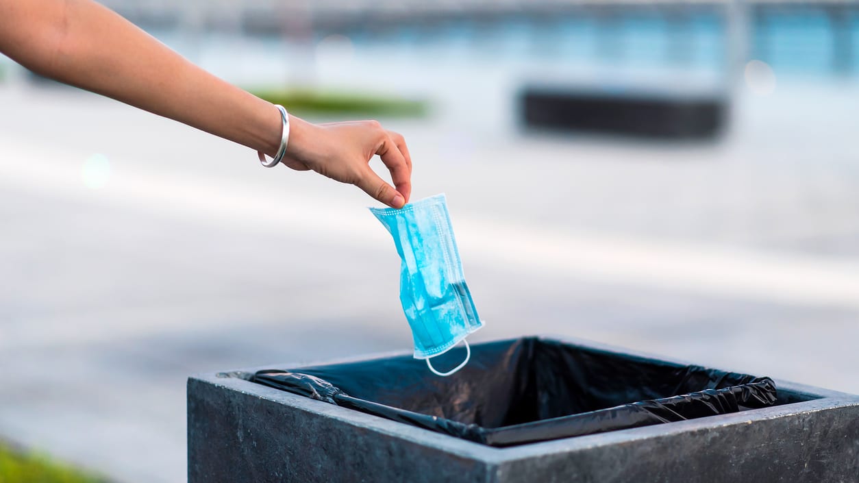 A person putting a blue face mask into a trash can.