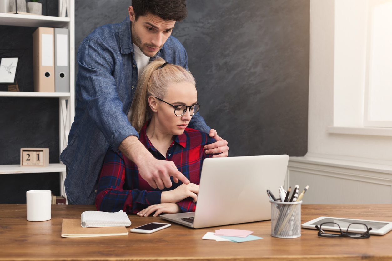 A male employee hovering over a female employee at her computer, gripping her left shoulder.