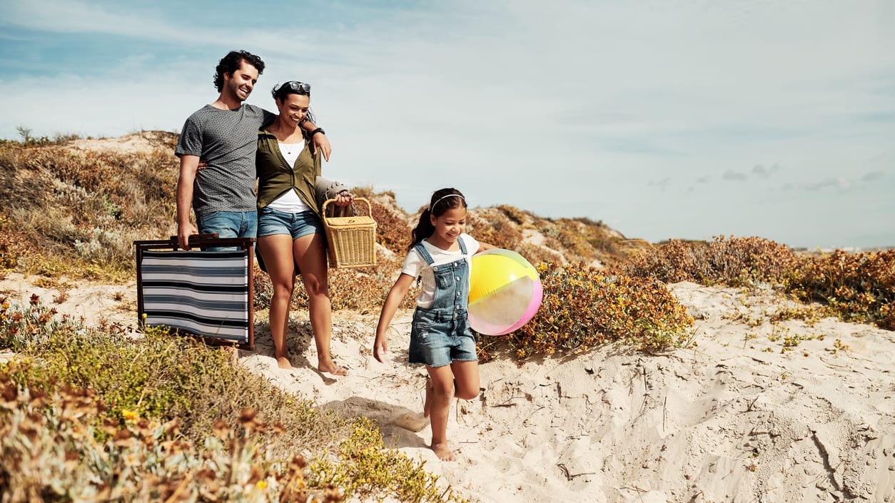 A family is walking down a sand dune with suitcases and a beach ball.