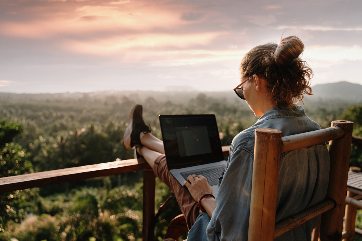 woman sitting on balcony outdoors overlooking beautiful scene outside while looking at her laptop