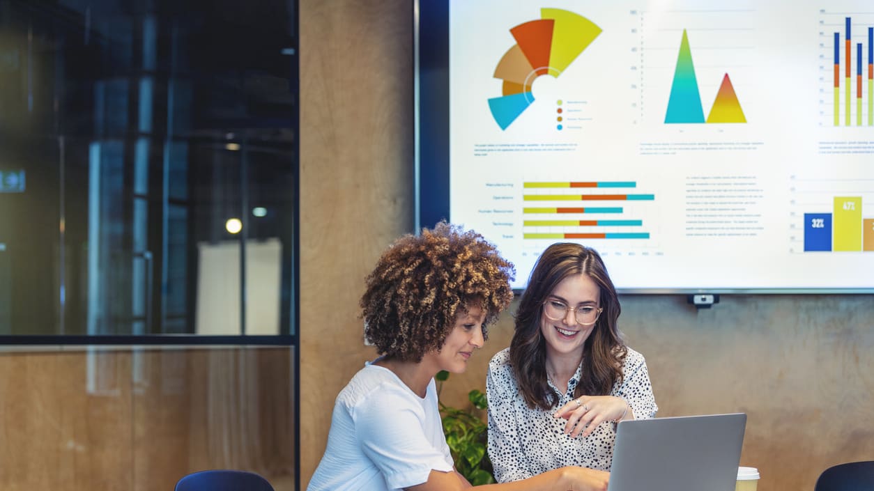 Two women working on a laptop in a conference room.