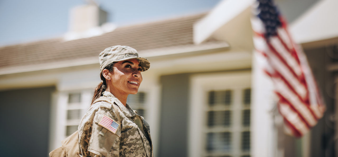 1428410222 Happy female soldier looking away with a smile while standing outside her house with her bag. American servicewoman coming back home after serving her country in the military.