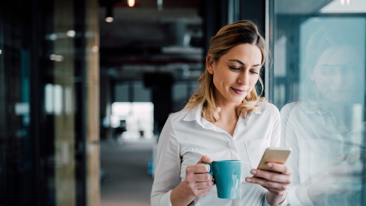 A business woman holding a cup of coffee while looking at her phone.