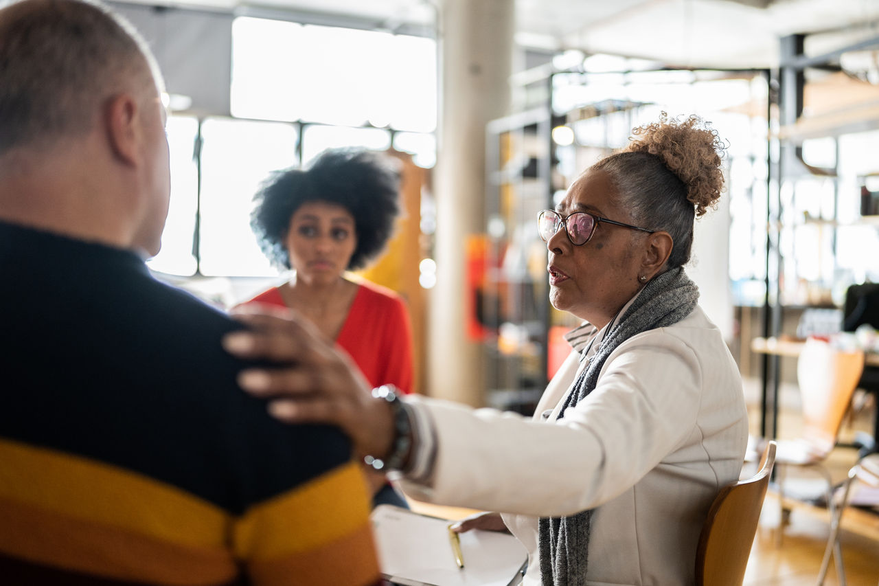 Older Black woman places hand on shoulder of man whose back is to the camera as younger Black woman looks on